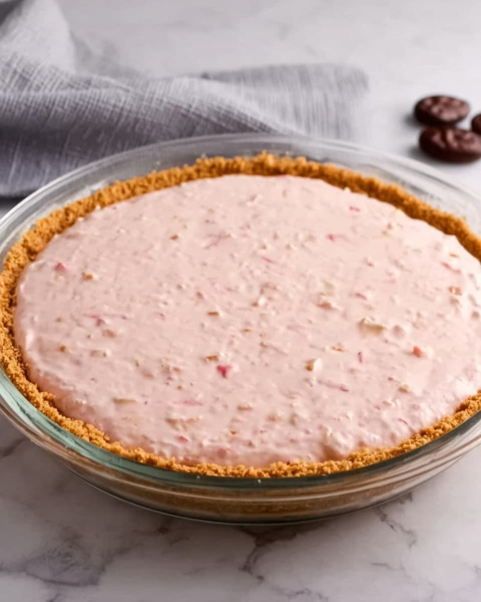 A round pie in a clear glass dish with a thick golden-brown crust holding a smooth, light pink filling with small bits of fruit or nuts mixed inside. The pie sits on a white marbled surface with a soft gray cloth in the background, and a few dark nuts are placed near the dish. The texture of the filling looks creamy and evenly spread, filling the crust fully up to the edges. photo taken with an iphone --ar 4:5 --v 7