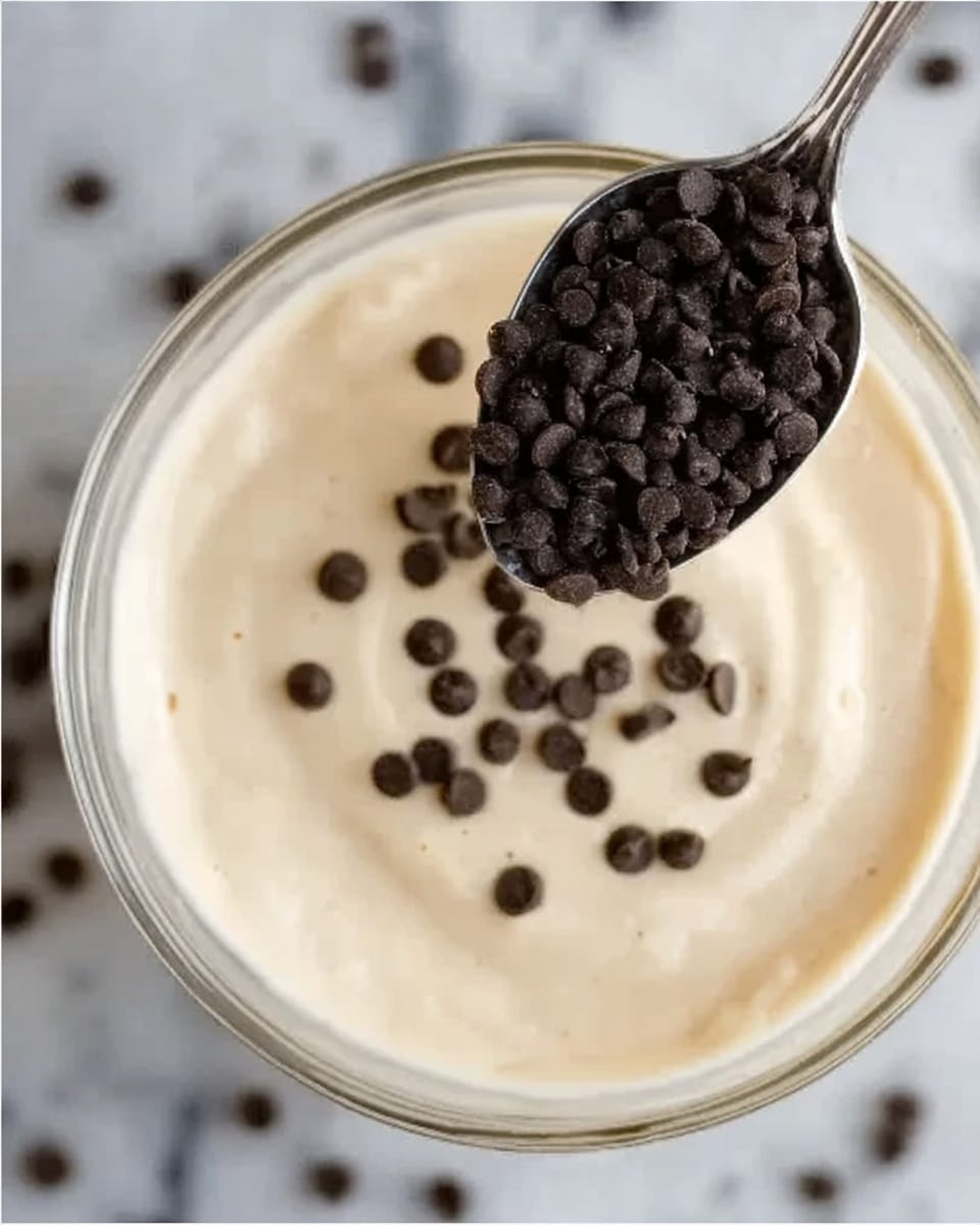 The image shows a close-up of a clear glass bowl filled with a smooth, light beige cream. On top of the cream, small dark brown chocolate chips are scattered. Above the bowl, a silver spoon holds a scoop of more chocolate chips, positioned to drop into the bowl. The background is a white marbled surface. photo taken with an iphone --ar 4:5 --v 7