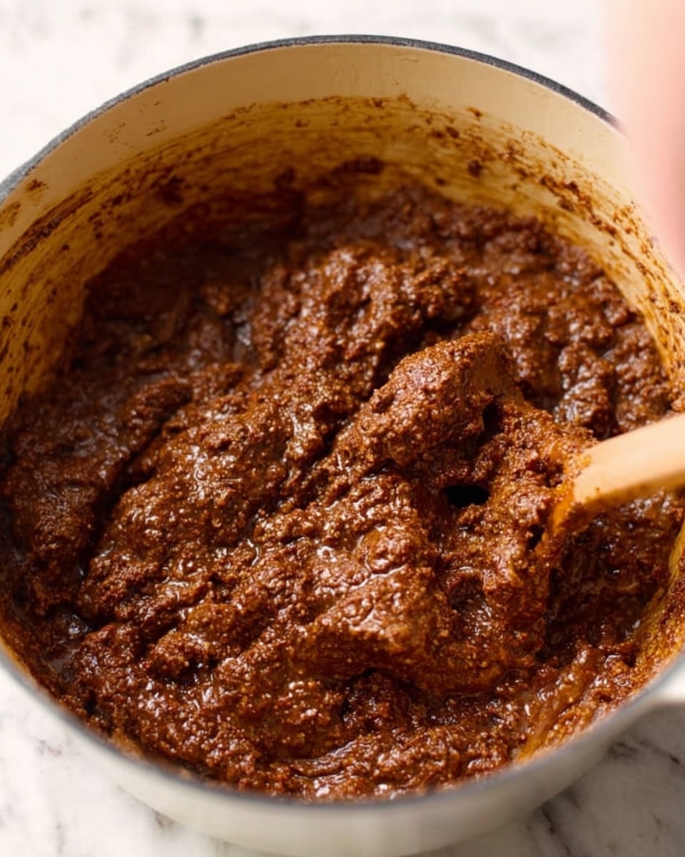 The image shows a close-up of a thick, dark brown meat mixture with a chunky and moist texture, spread inside a white pot. The surface of the meat looks soft with visible small bits and a little oily shine. The background is a white marbled texture. A blurred woman’s hand is stirring the mixture with a wooden spoon from the top right corner. photo taken with an iphone --ar 4:5 --v 7