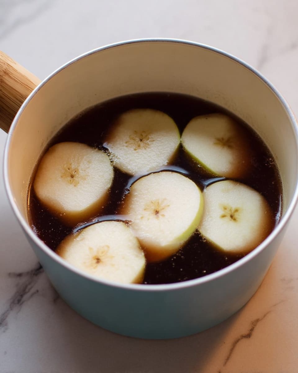 A white pot with a light blue outside edge holds a dark brown liquid filled almost to the top. Floating on the surface are six thick, pale cream slices of peeled apple, each piece showing smooth edges and a slightly sunken center. The pot has a light wooden handle on the left side, and all items sit on a white marbled surface. photo taken with an iphone --ar 4:5 --v 7