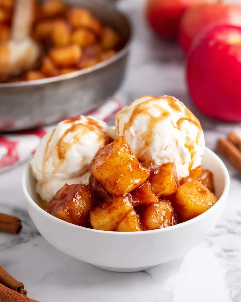 The image shows a white bowl filled with two scoops of creamy vanilla ice cream, topped with warm, golden-brown apple pieces coated in a shiny cinnamon glaze. The apple pieces are soft and chunky, sitting neatly on top of the ice cream, slightly melting into it. The bowl is placed on a white marbled surface, with a skillet of more glazed apple pieces blurred in the background along with red apples and cinnamon sticks partially visible. The overall look is cozy and inviting, with warm amber and cream colors standing out. photo taken with an iphone --ar 4:5 --v 7