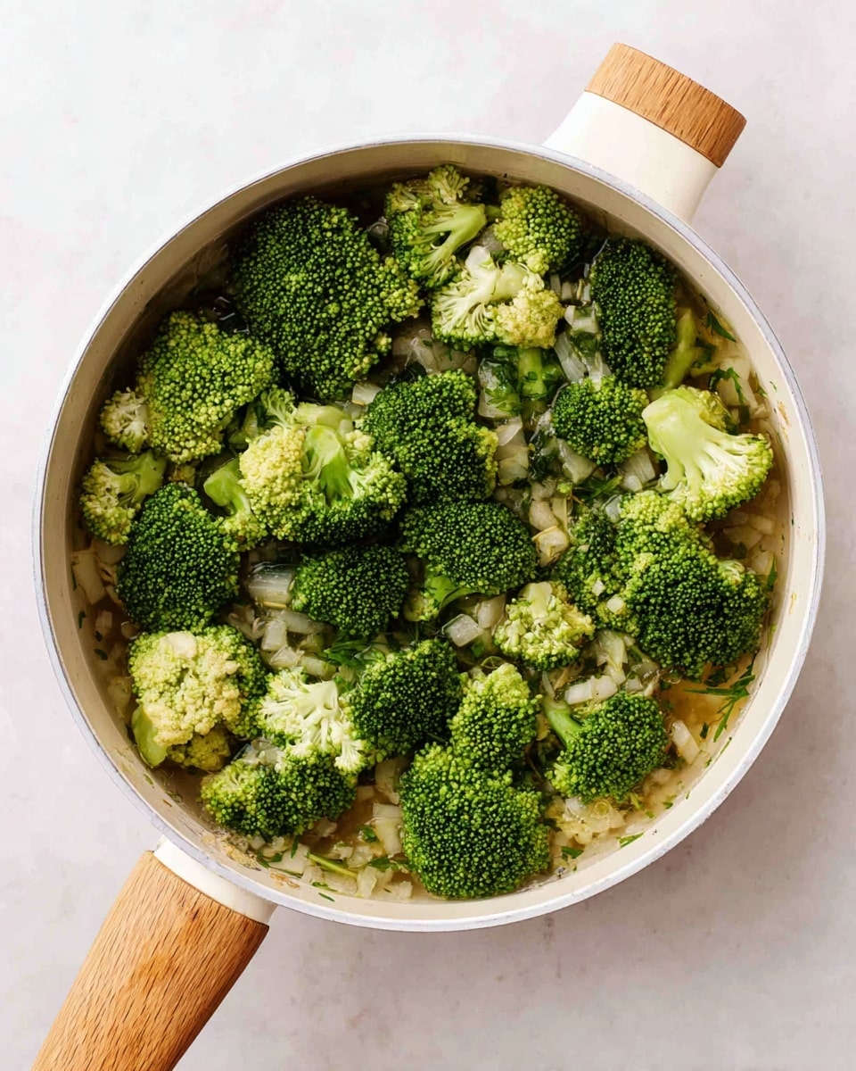 A white pan with a light wooden handle is filled with bright green broccoli florets on top. Underneath the broccoli, there are small pieces of chopped onion and herbs mixed in a clear broth. The broccoli pieces have a slightly rough texture with tiny flower buds, and the onion bits are translucent and soft, resting at the bottom. The pan is placed on a white marbled surface, creating a clean and fresh look. photo taken with an iphone --ar 4:5 --v 7
