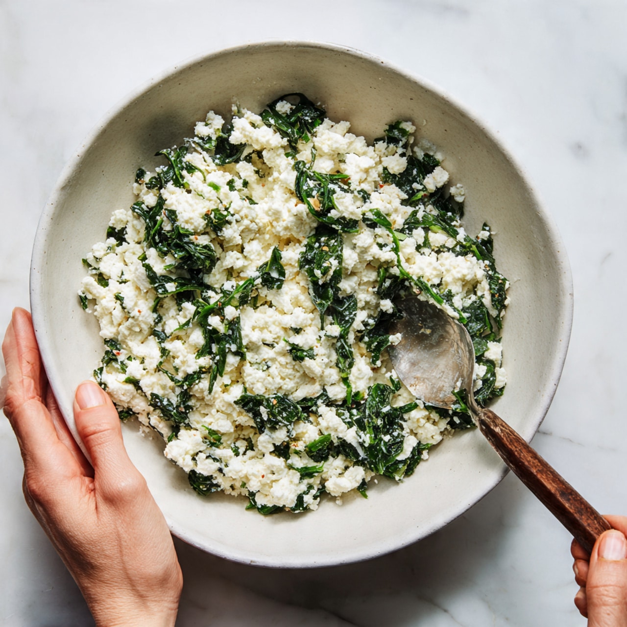 A white bowl filled with a mix of white, soft, crumbly cheese and chopped green leafy vegetables, creating a textured and uneven surface with small clumps of both ingredients across the bowl. One woman's hand holds the bowl steady, while another woman's hand uses a spoon to stir the mixture, showing movement and interaction with the food. The background is a white marbled texture that softly contrasts with the white bowl and the dark green and white mixture inside. Photo taken with an iphone --ar 4:5 --v 7