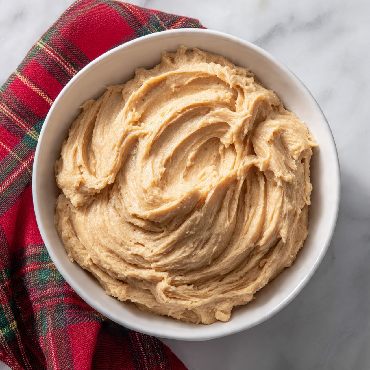 A white bowl filled with light brown cookie dough that looks smooth but thick with some visible soft peaks and swirls from mixing, sitting on a white marbled surface. Next to the bowl is a red and green plaid cloth adding a splash of color to the scene. The dough fills most of the bowl and has a creamy texture with slight indentations from stirring visible on top photo taken with an iphone --ar 4:5 --v 7