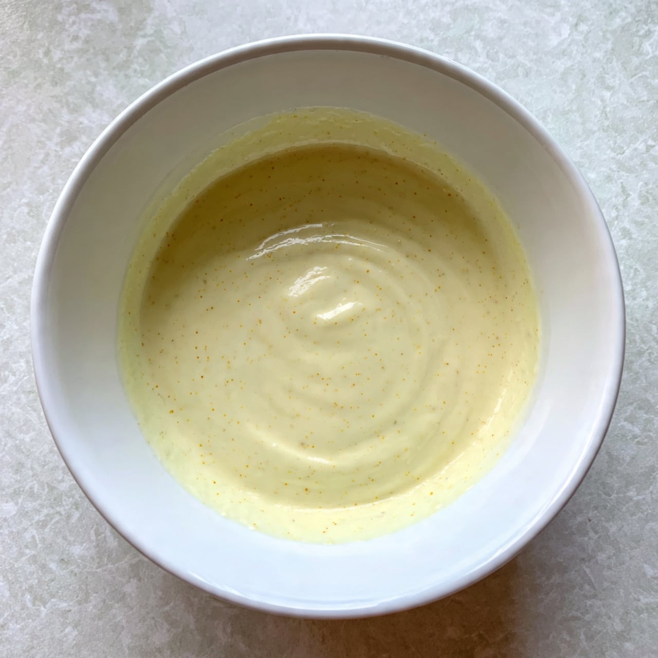 A round white bowl filled with smooth, creamy batter that is pale yellow with a hint of speckled texture, evenly spread inside the bowl with gentle swirling patterns on the surface. The bowl sits on a white marbled textured surface. photo taken with an iphone --ar 4:5 --v 7