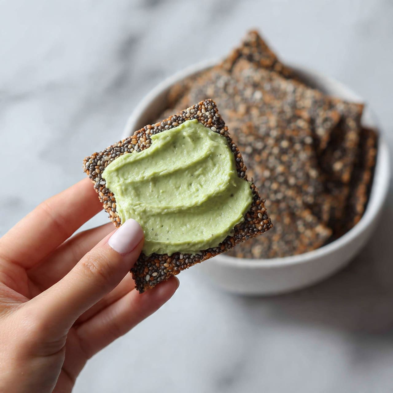 A close-up image of a woman's hand holding a square beige cracker with a rough texture, partially covered with a thick, light green creamy spread, showing small lumps. In the background, there is a blurred white bowl filled with similar beige crackers resting on a white marbled surface. The lighting is soft and natural, highlighting the textures of the cracker and the spread. Photo taken with an iphone --ar 4:5 --v 7