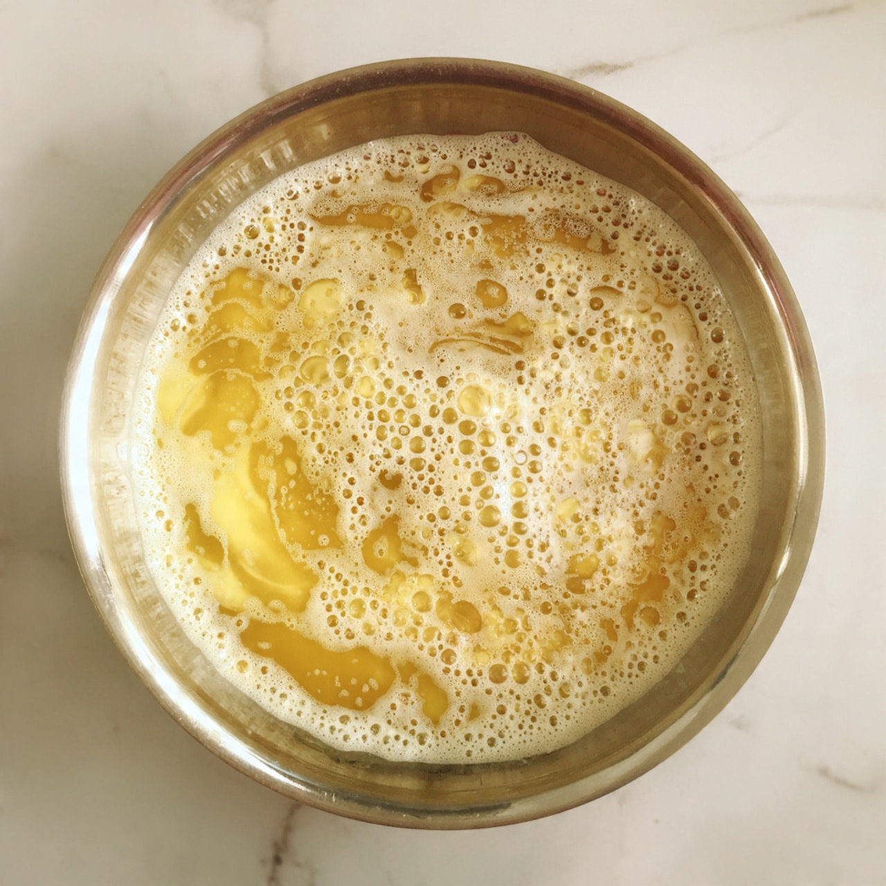 A close-up top view of the inside of a shiny stainless steel pan with some melted butter heating up. The butter is golden yellow with white foamy bubbles forming unevenly on the surface in swirling patterns. The pan is placed on a white marbled surface. Photo taken with an iphone --ar 4:5 --v 7