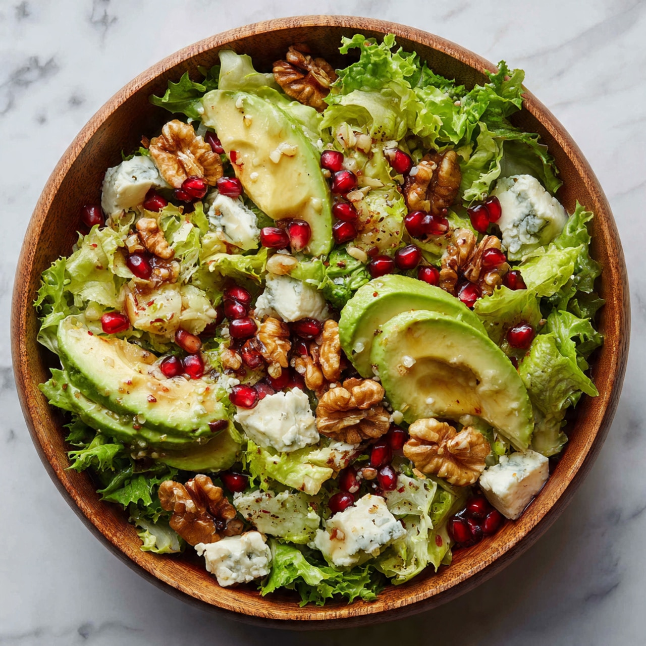 A wooden bowl filled with a colorful salad sits on a white marbled surface. The bottom layer is made of green leafy lettuce with frilly edges, spread evenly throughout. Scattered on top are bright red pomegranate seeds adding small bursts of color. Large, creamy avocado slices with smooth, light green flesh sit among the lettuce. There are pieces of white cheese with blue veins, crumbly in texture, layered generously across the salad. Several golden brown walnuts and roasted seeds bring a crunchy texture, lightly coated and sprinkled evenly. The salad has a shiny drizzle of dressing visible on top, enhancing the freshness of the ingredients. photo taken with an iphone --ar 4:5 --v 7