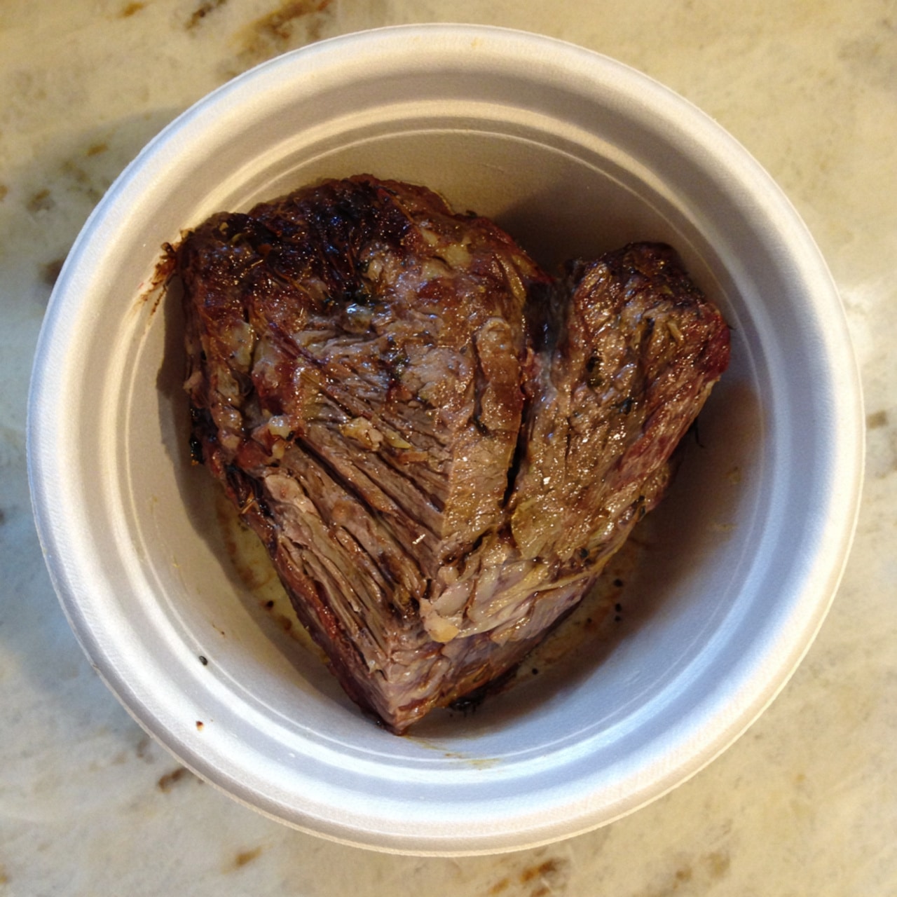 A single large piece of cooked meat with a browned, textured surface sits inside a white round bowl, showing different shades of brown with some darker cooked spots and visible lines of the meat grain. The bowl is placed on a white marbled surface that adds a clean, light background to the image. Photo taken with an iphone --ar 4:5 --v 7