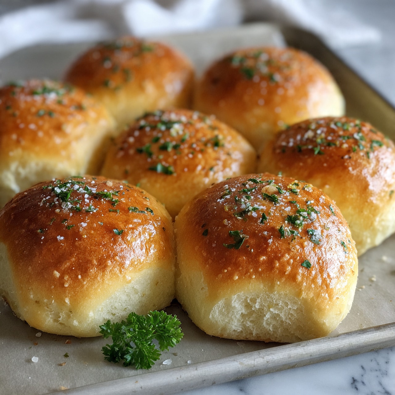 A close-up view of several small, round golden bread rolls placed closely together on a light gray baking sheet. Each roll has a shiny, browned top sprinkled with green parsley flakes and small granules of salt, giving a textured look. The bread surface shows a soft, smooth texture with subtle browning and hints of a brushed butter glaze. Some dark pepper specks are lightly scattered on the rolls' tops, and a small sprig of fresh parsley rests on the bottom left corner of the baking sheet. The background is a white marbled texture. photo taken with an iphone --ar 4:5 --v 7