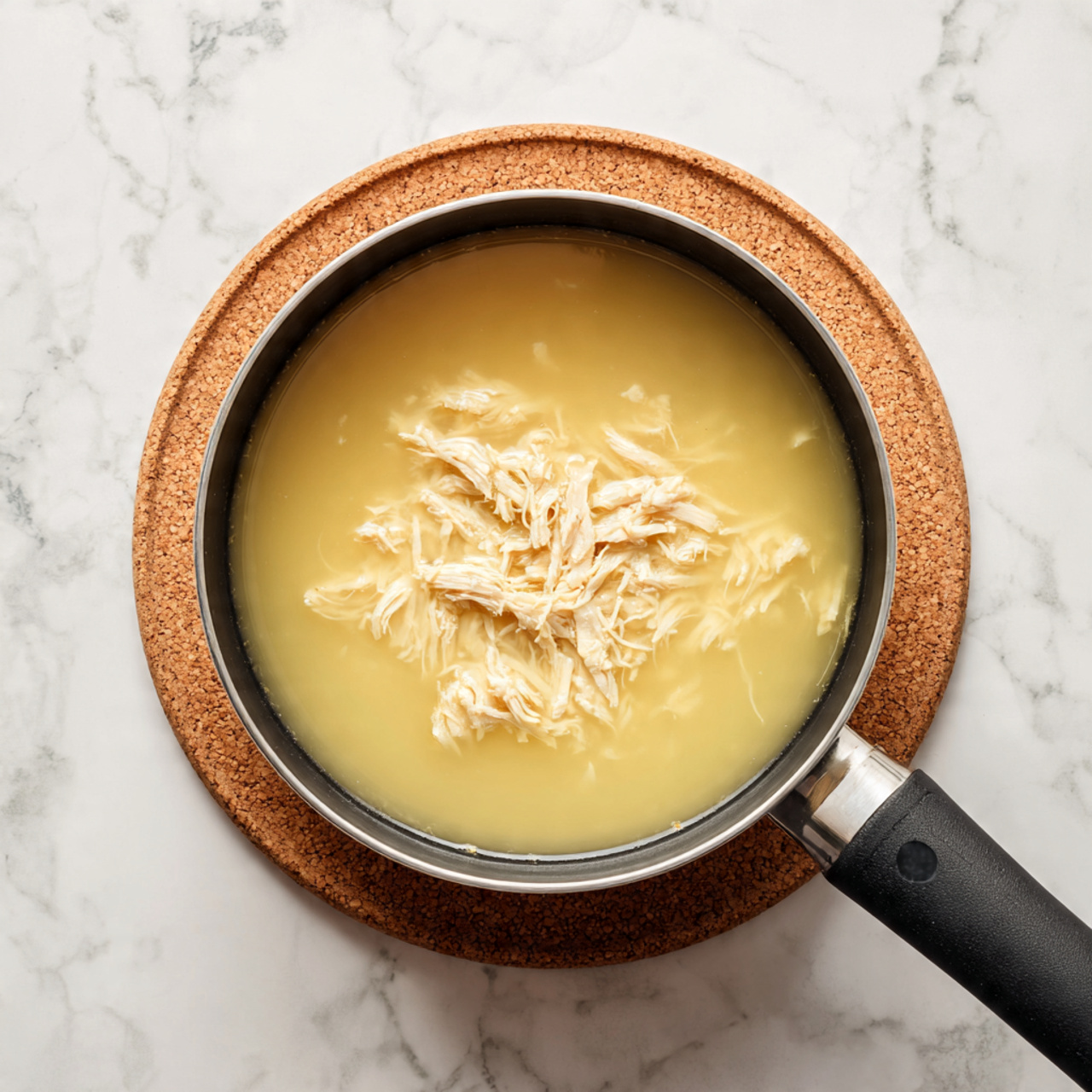 The image shows a black saucepan filled with light yellow soup or broth, with shredded light brown meat piled in the center of the liquid. The saucepan rests on a cork mat, which is placed on a white marbled surface. The broth looks smooth and slightly thick, while the meat strands are soft and spread evenly in the middle. photo taken with an iphone --ar 4:5 --v 7