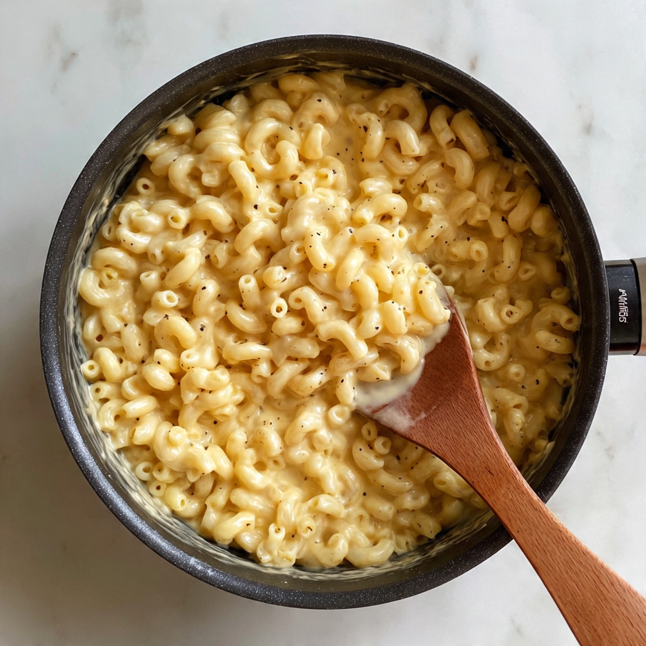 The image shows a close-up of a black pot filled with creamy macaroni pasta. The pasta is small, curved, and coated evenly in a pale yellow cheese sauce with a smooth, thick texture. A wooden spoon is stirring the macaroni, showing the sauce's creamy consistency, with small black pepper specks visible on top. The background surface is a white marbled texture. Photo taken with an iphone --ar 4:5 --v 7