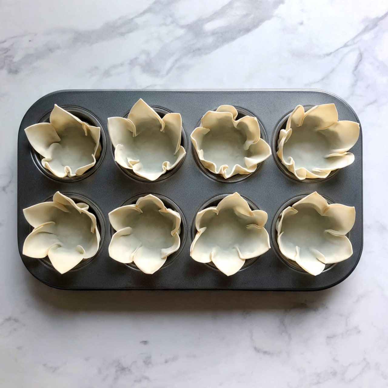 A silver muffin tin with twelve cups, each lined with a single layer of light tan, thin wonton wrappers that are folded and slightly crinkled, creating small pockets with four points sticking up like petals. The tin rests on a white marbled surface, and there are tiny droplets of water scattered on the tin, giving it a fresh look. The background is blurred and neutral, focusing attention on the empty wonton cups. photo taken with an iphone --ar 4:5 --v 7