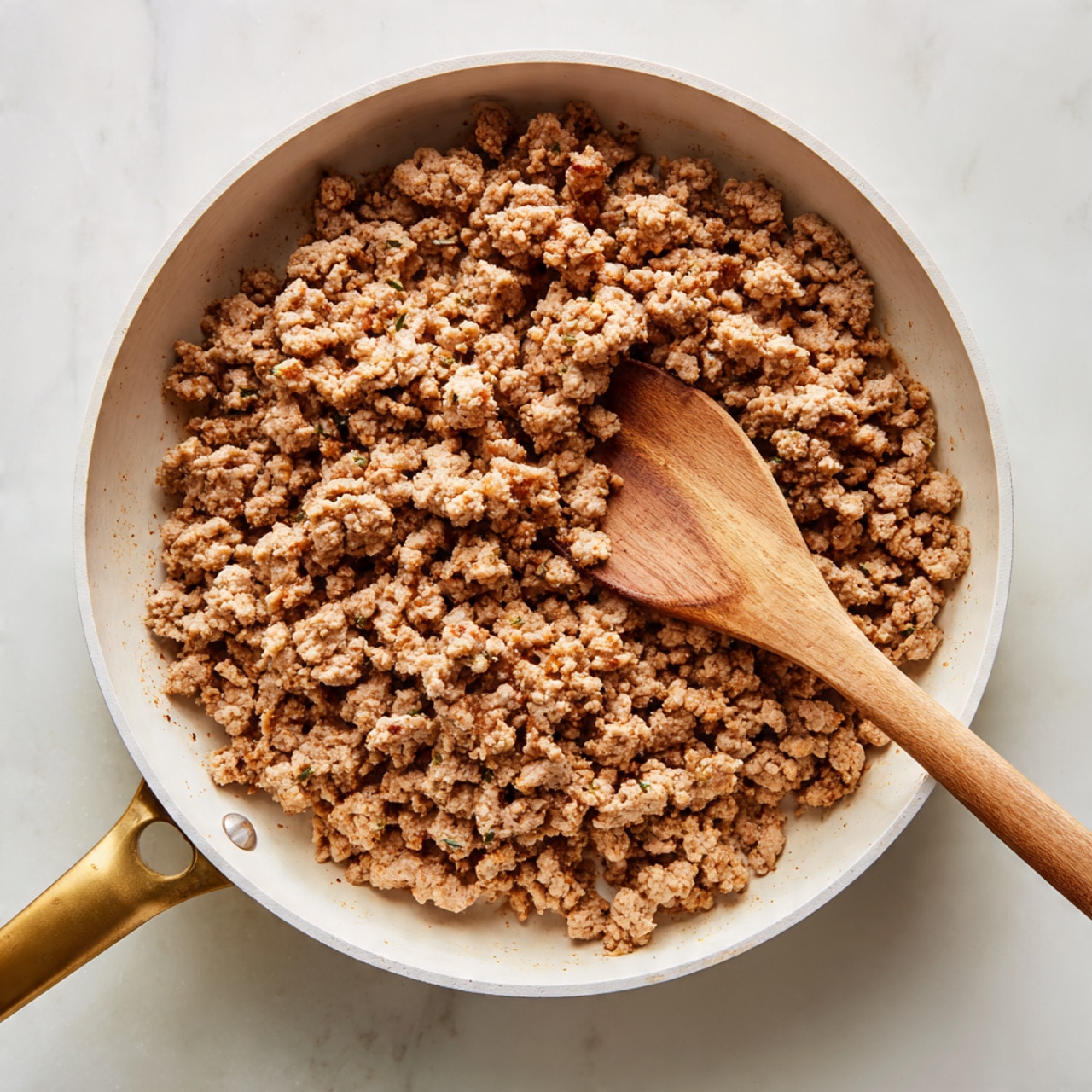 In a white pan with a golden handle, there is one layer of cooked ground meat spread evenly across the surface. The meat is crumbly with a soft brown color and slight hints of fat and juices mixed in. A wooden spoon with a smooth texture rests on the right side of the pan, partially covered by the meat. The background is a white marbled texture. photo taken with an iphone --ar 4:5 --v 7