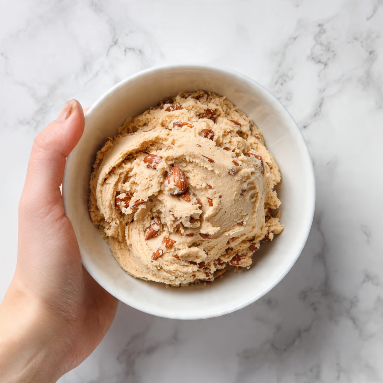 A white bowl filled with light brown cookie dough that has a soft, creamy texture. Mixed inside the dough are whole and broken almonds, scattered evenly throughout. The bowl sits on a white marbled surface. A woman's hand is seen at the top left corner of the image, gently resting near the bowl. Photo taken with an iphone --ar 4:5 --v 7