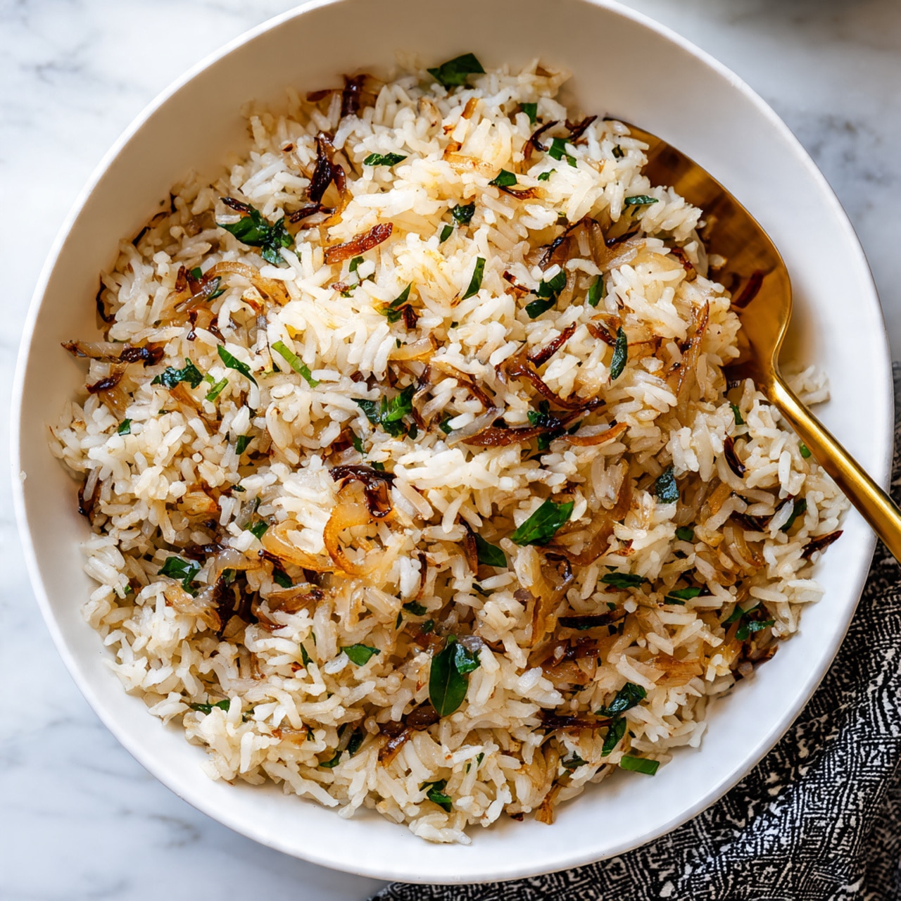 A close-up view of a white bowl filled with cooked rice mixed with small diced brown onions and sprinkled with small green herb leaves. The rice grains are fluffy and light brown with some caramelized onion pieces scattered throughout. A gold-colored spoon is partially buried in the rice, showing the texture of individual rice grains and onions. The bowl sits on a white marbled surface with a glimpse of a cloth napkin with black and white patterns at the edge of the frame. photo taken with an iphone --ar 4:5 --v 7