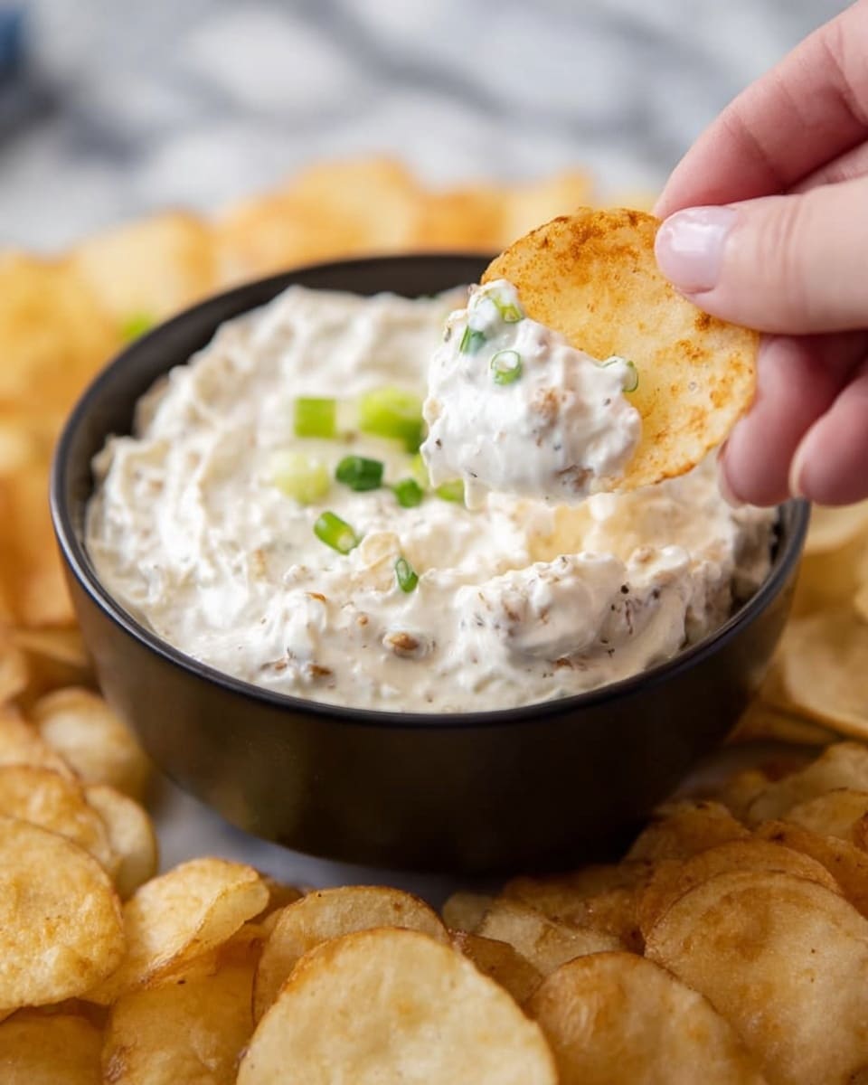 A close-up of a woman's hand holding a round, light golden potato chip covered in thick, creamy white dip with small brown bits. The chip is dipped into a black bowl filled with the same creamy dip, garnished with small green onion pieces on top. Surrounding the bowl is a spread of similar round potato chips. The surface beneath is white marble. photo taken with an iphone --ar 4:5 --v 7