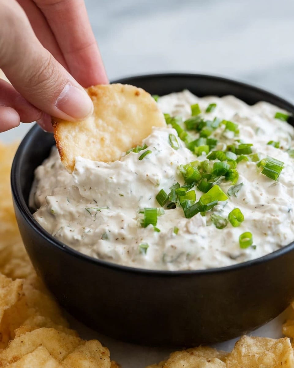 A close-up of a black bowl filled with thick, white creamy dip with small chunks mixed inside. On top of the dip are scattered bright green chopped scallions. A woman's hand is holding a light beige round chip dipping into the bowl, coated with the creamy dip. The bowl is placed on a white marbled surface with some similar chips around the bowl. photo taken with an iphone --ar 4:5 --v 7