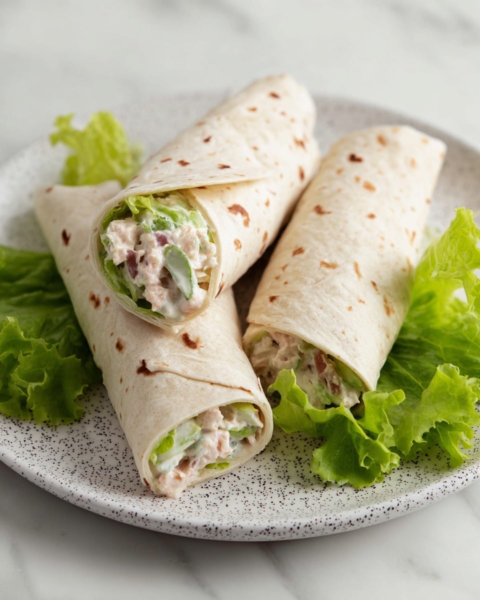 The image shows three small tortilla wraps placed together on a white plate with dark speckles, set on a white marbled surface. Each wrap has a light, beige color with visible small seeds or grains scattered throughout the soft tortilla. The wraps are filled with layers of creamy white sauce mixed with pieces of green lettuce and small bits of what looks like light pink tuna or chicken salad visible at the open ends. Around the wraps, there are a few fresh green lettuce leaves for garnish. The photo taken with an iphone --ar 4:5 --v 7