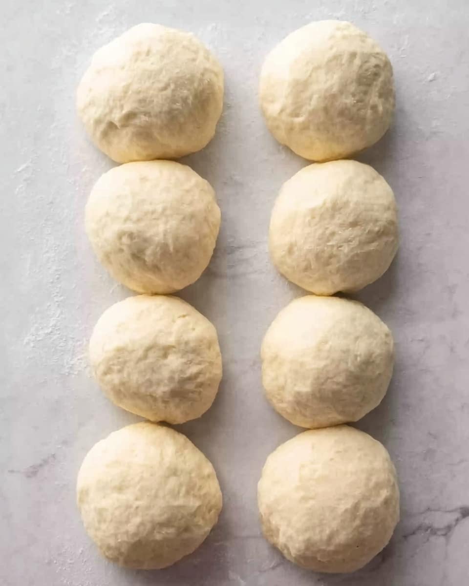 The image shows eight dough balls arranged in two even columns of four, placed on a white marbled surface. Each ball is light beige in color with a soft, slightly irregular texture suggesting they have been freshly formed and are ready for baking or cooking. The dough balls are smooth on top but show faint dimples and creases, giving them a homemade look. The spacing is neat, and the surface beneath them has subtle grayish veins typical of white marble. photo taken with an iphone --ar 4:5 --v 7