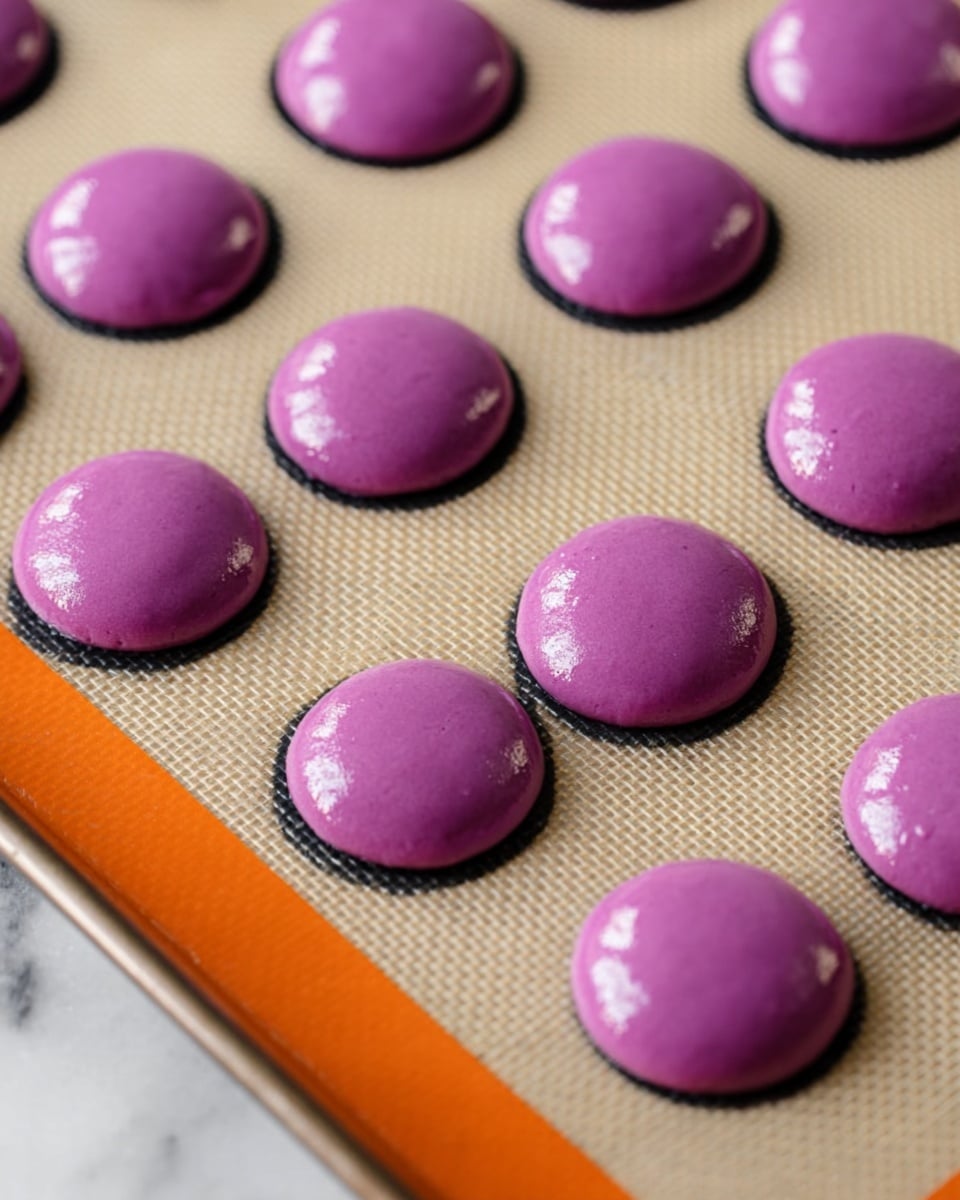 The image shows a baking sheet lined with a beige silicone mat that has black circular guides. On the mat, there are evenly spaced, smooth, glossy, round dollops of bright purple batter, arranged in rows. Each piece is slightly domed and looks soft with a shiny surface texture. The baking sheet edges are visible with a touch of orange color at the top and bottom. The background is a white marbled surface. photo taken with an iphone --ar 4:5 --v 7