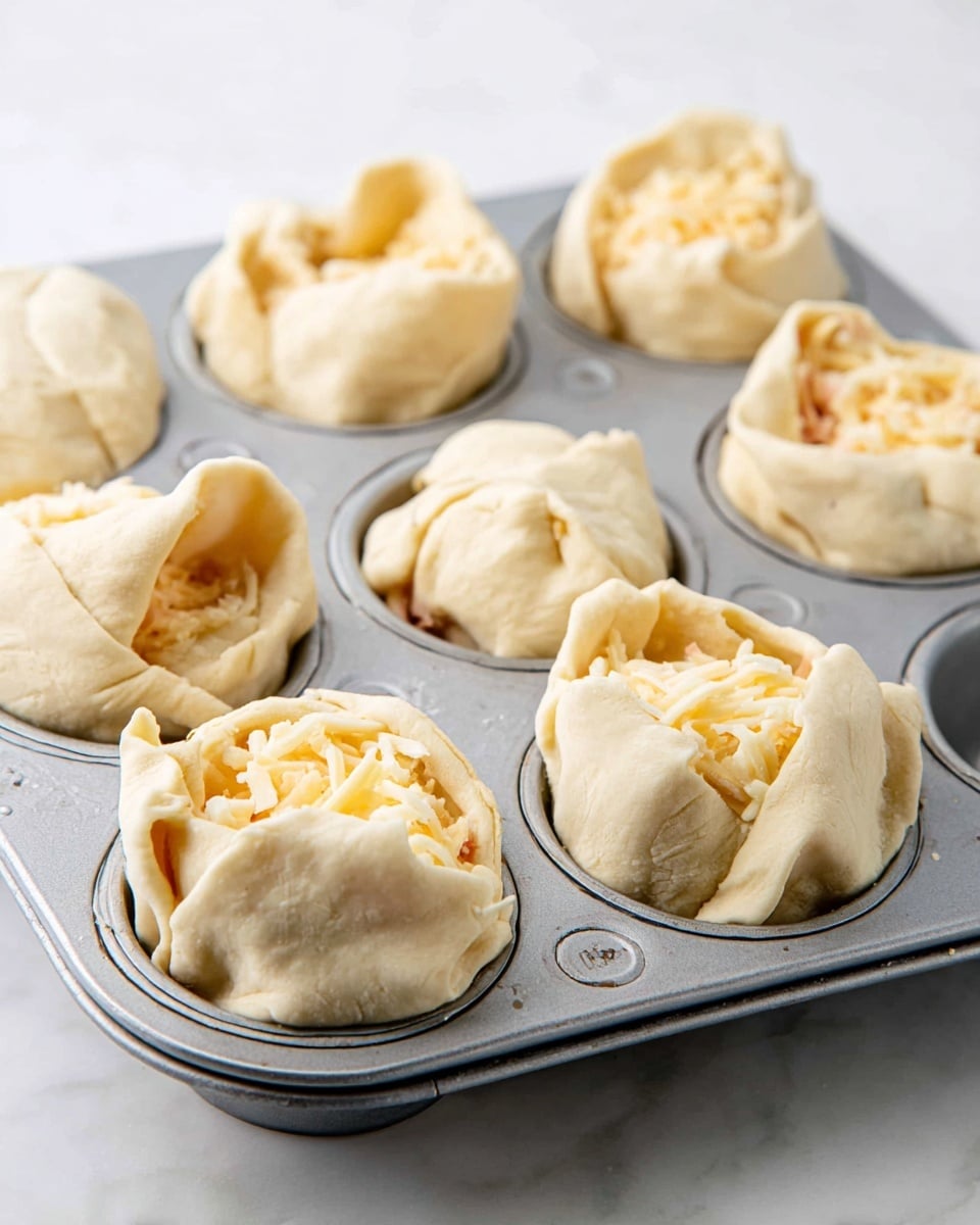 The image shows six uncooked dough pockets in a metal muffin tray, each filled with a mixture of shredded yellow and white cheese visible through the slightly wrapped pale dough. Each dough pocket is folded unevenly, creating soft layers of light beige dough with some cheese peeking out from the folds. The metal muffin tray has a smooth, round indented pattern inside each cup. The background is a white marbled surface, giving a clean and bright look. photo taken with an iphone --ar 4:5 --v 7
