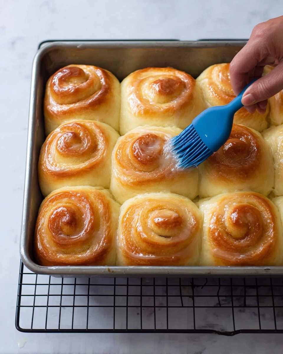 A metal baking tray holds twelve golden brown rolls with a soft, fluffy texture, arranged closely in a 3 by 4 grid. Each roll has a spiral shape, showing the layers of dough with a shiny, slightly sticky surface from a glaze being brushed on by a woman's hand holding a blue silicone brush. The tray is placed on a black wire cooling rack on top of a white marbled surface. The rolls' outer edges have a light brown crispness while the inner parts remain pale and soft. Photo taken with an iphone --ar 4:5 --v 7
