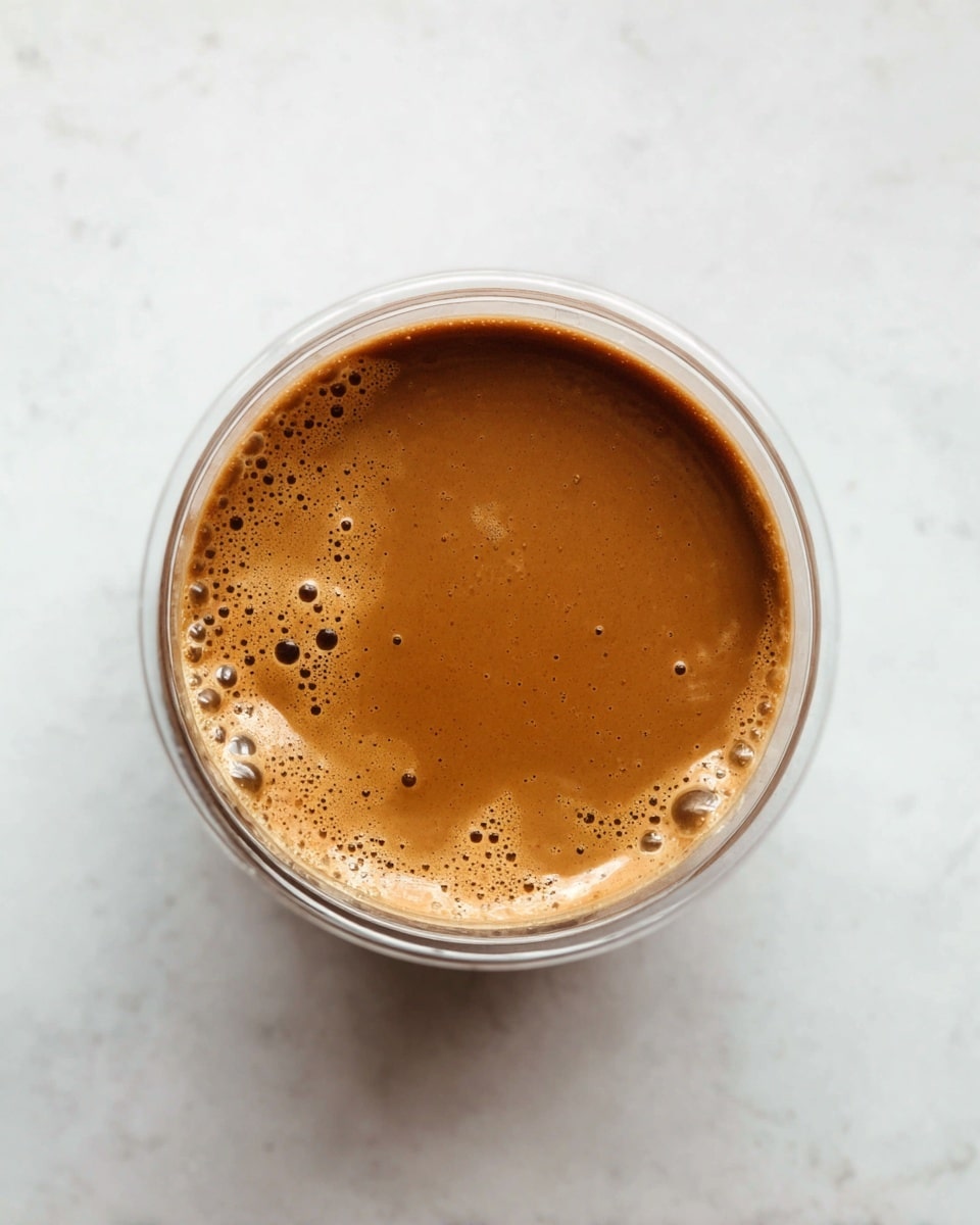 A top view of a single round clear container filled with smooth, thick, light brown sauce that has small bubbles and a slightly shiny surface. The container sits on a white marbled texture. photo taken with an iphone --ar 4:5 --v 7