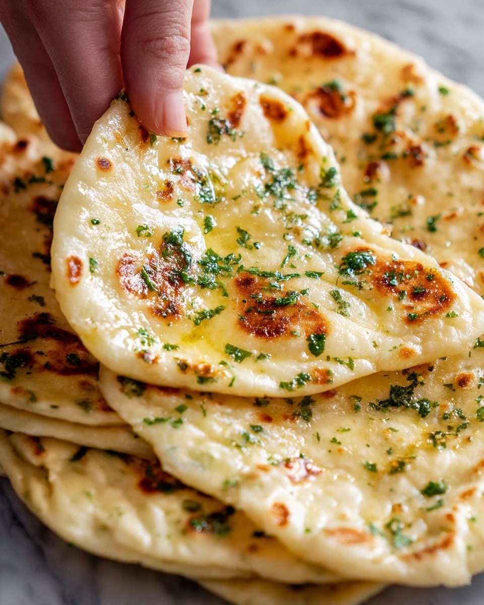 The image shows a close-up of flatbread being lifted by a woman's hand. The flatbread has a light golden color with some darker toasted spots and is sprinkled with chopped green herbs on top. The texture looks soft and slightly puffy, with some melted butter or oil giving it a shiny surface. Multiple layers of the flatbread are stacked under the piece being lifted, all on a white marbled surface. photo taken with an iphone --ar 4:5 --v 7