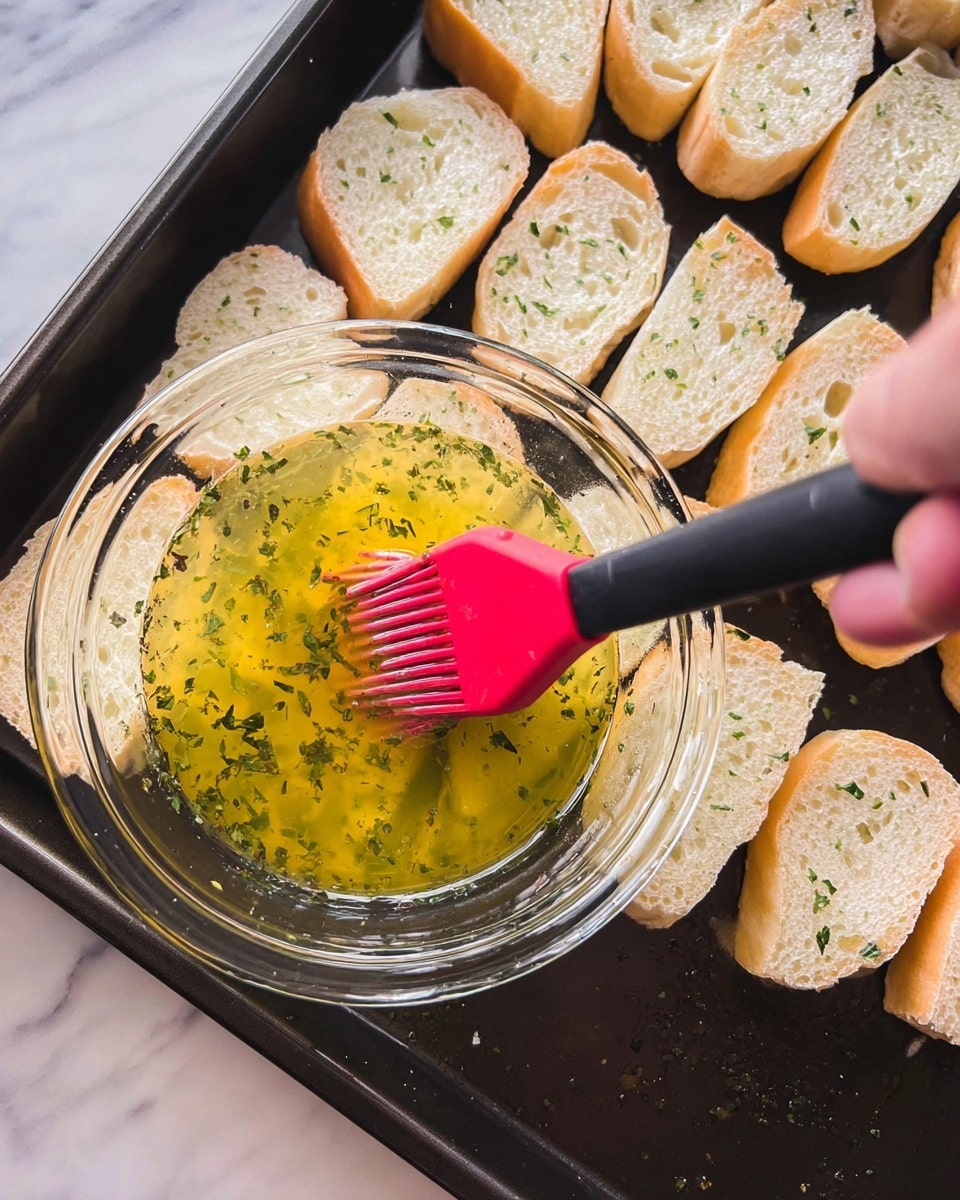 A close-up view of a black baking tray filled with three rows of small, white bread slices with light golden crusts at the edges. In the foreground, a woman's hand holds a clear glass bowl containing a yellow, oily mixture with bits of green herbs. A black and red silicone brush rests in the bowl, partially covered in the mixture. The scene is set on a white marbled textured surface. photo taken with an iphone --ar 4:5 --v 7