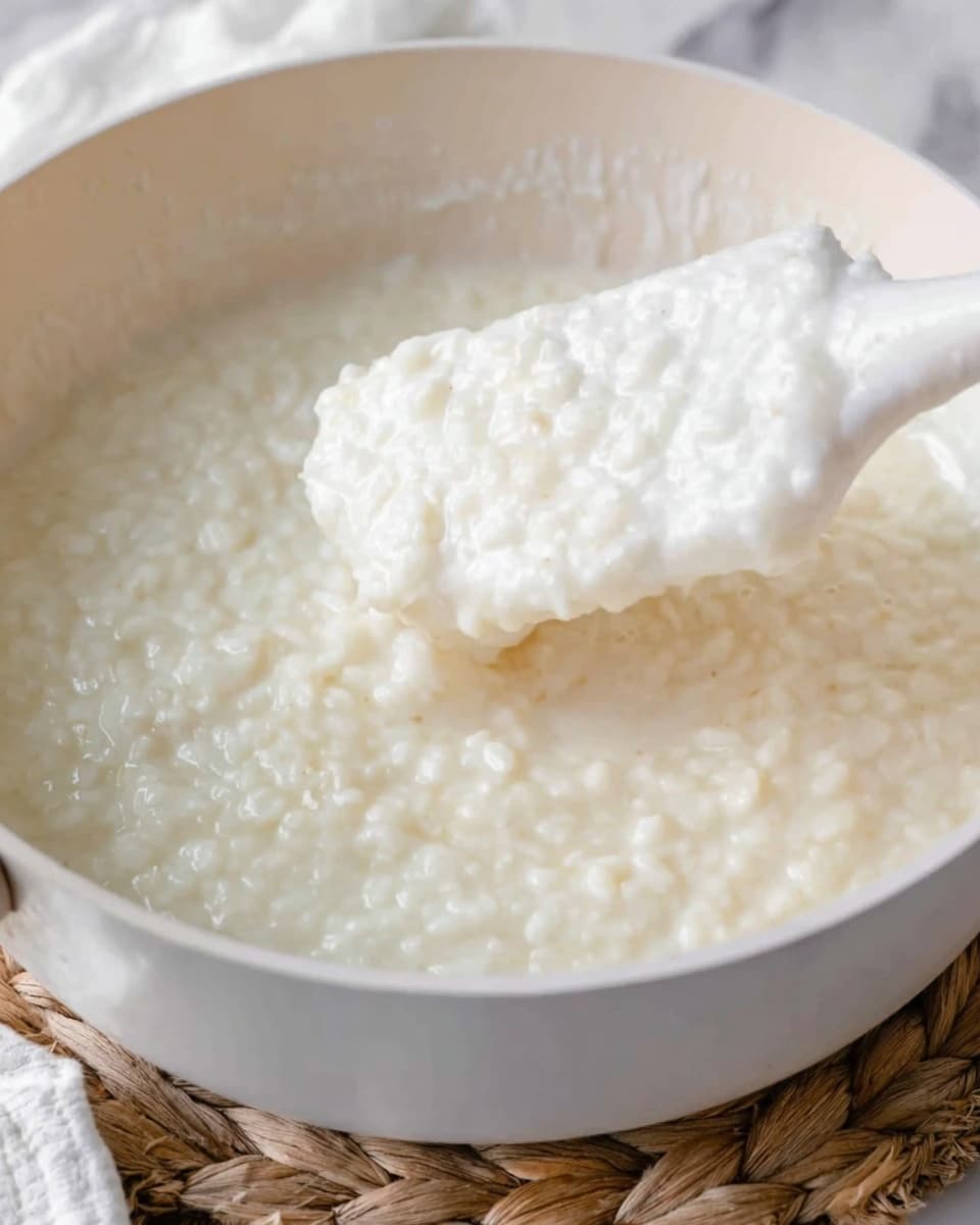 The image shows a close-up of a creamy, white rice pudding with a thick and lumpy texture in a white pan. A white spatula is lifting some of the pudding, showing its soft and slightly sticky consistency, with small round grains visible throughout. The background features a white marbled surface and part of a woven mat under the pan, adding a natural touch. photo taken with an iphone --ar 4:5 --v 7