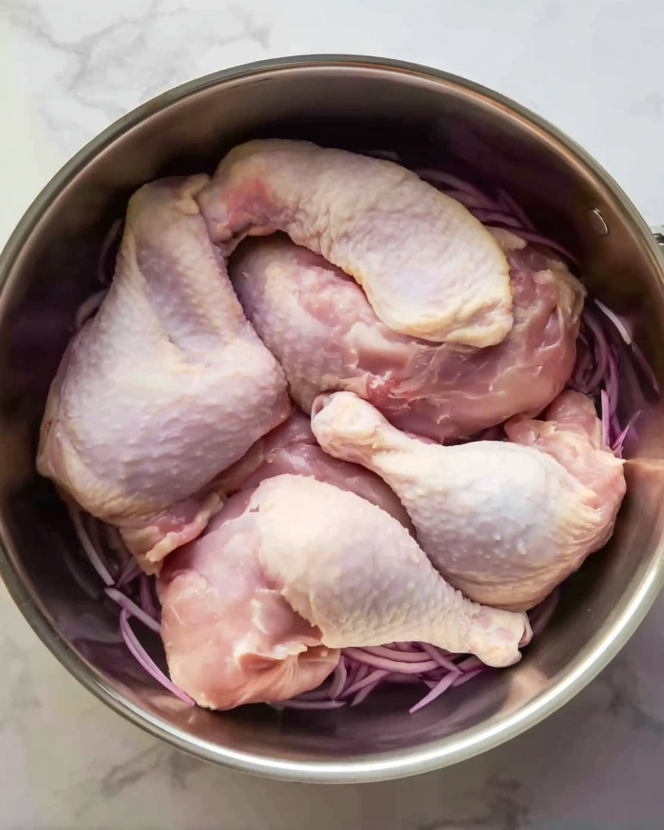 Three raw chicken leg quarters with pale pink skin and some white fat are placed on top of thinly sliced purple onion layers at the bottom of a round metal cooking pot. The chicken legs are arranged close together, with the drumsticks pointing to different sides. The pot has a shiny silver metal inside surface, and the onions beneath the chicken add some purple color and texture. The background is a white marbled texture. Photo taken with an iphone --ar 4:5 --v 7
