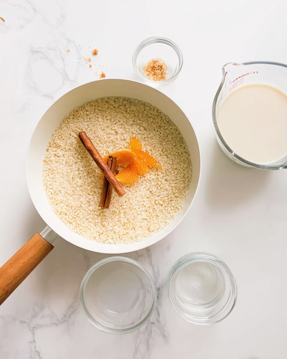 A white pan with a wooden handle sits on a white marbled surface, filled with a layer of small rice grains in liquid, topped with a cinnamon stick and orange peel strips placed in the center. Around the pan are clear glass bowls and a clear measuring cup filled with milk, all arranged neatly. The colors are soft with white, beige, and orange tones against the bright, clean background. photo taken with an iphone --ar 4:5 --v 7