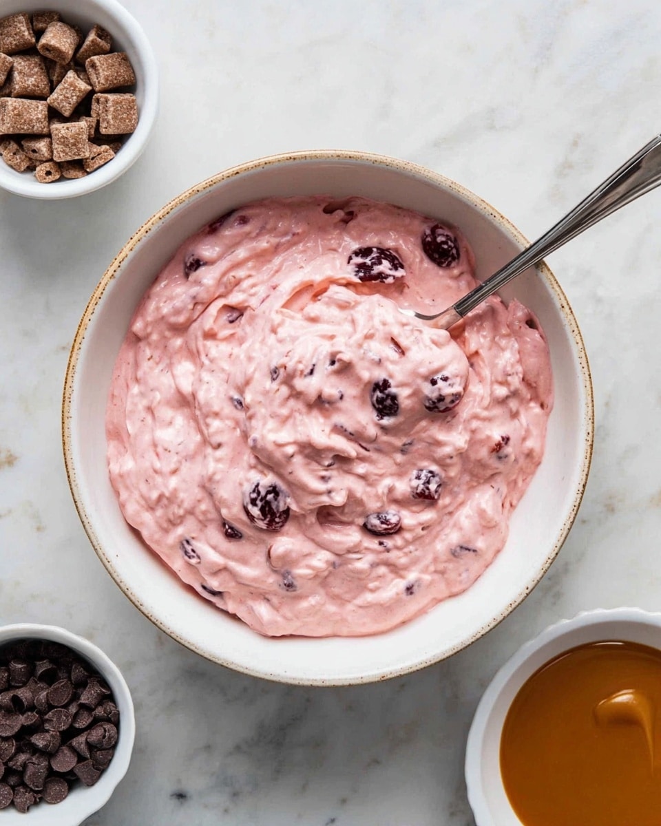 The image shows a bowl filled with a thick pink creamy mixture that has visible dark cherry-like pieces mixed in, giving it a textured look with small lumps and fruit chunks spread throughout. The bowl is white with a light brown rim and spots, and inside it, there is a silver spoon partially dipped in the pink mixture. Surrounding the bowl on a white marbled surface, there are three small white bowls, one containing small brown square cereal pieces, another with dark chocolate chips, and the third holding a smooth caramel-colored sauce. The scene is bright and clean, giving a fresh and inviting feel. photo taken with an iphone --ar 4:5 --v 7