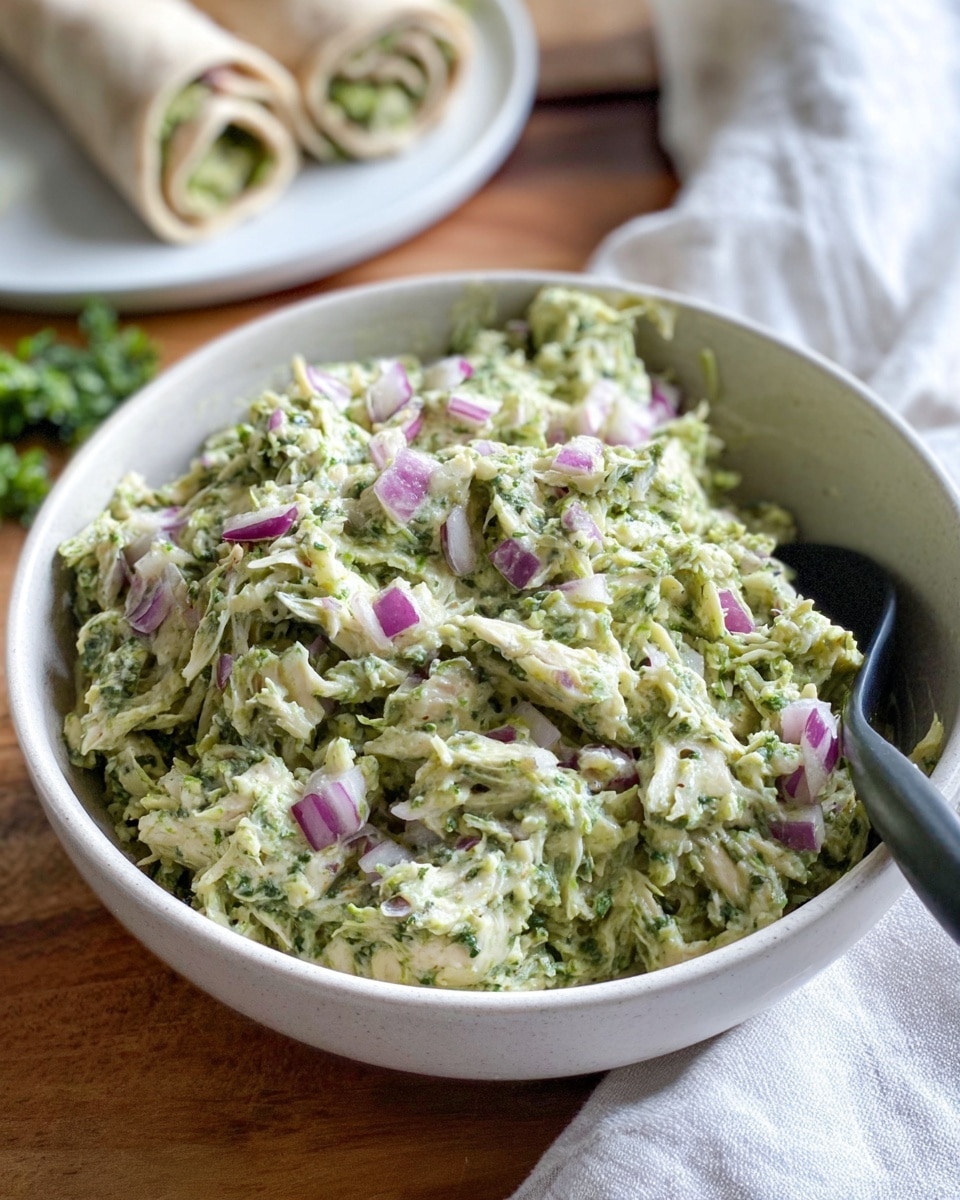 A close-up view of a white bowl filled with a green shredded mixture that looks creamy and herb-filled, topped with small pieces of chopped red onion adding a pop of purple and white. The texture is soft and fluffy, with visible strands and bits of the mixture well-mixed together. A black spoon rests inside the bowl on the right side. The bowl is placed on a wooden surface, with a white cloth and some green herbs out of focus in the background. In the distant background, there is a blurry white plate holding rolled wraps. photo taken with an iphone --ar 4:5 --v 7