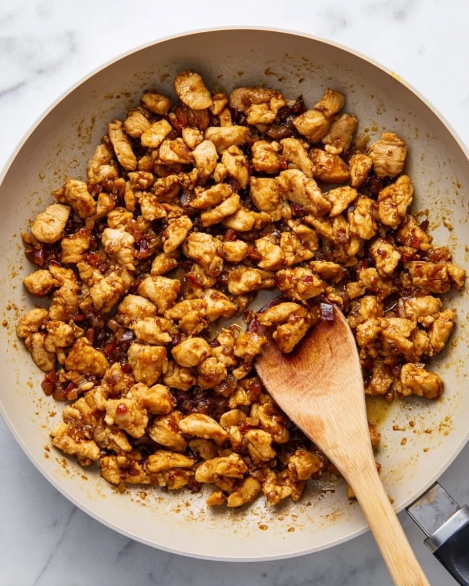 The image shows a white pan filled with small chunks of cooked meat that are golden brown and slightly crispy. The meat is spread evenly across the surface of the pan, with a wooden spoon resting in the middle, scooping a few pieces. The background is a white marbled surface, which contrasts with the warm colors of the meat and the pale color of the pan. photo taken with an iphone --ar 4:5 --v 7