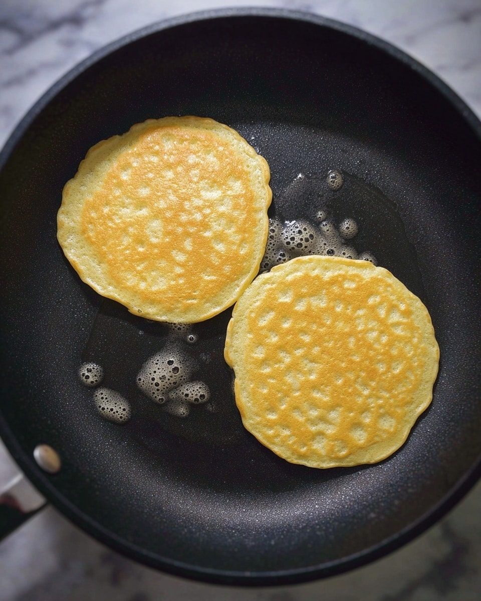Two round yellow pancakes cooking side by side in a black non-stick pan, each pancake showing a slightly bubbly and uneven surface texture with soft edges, surrounded by small glistening oil bubbles on the pan’s surface, all set against a white marbled texture background, photo taken with an iphone --ar 4:5 --v 7