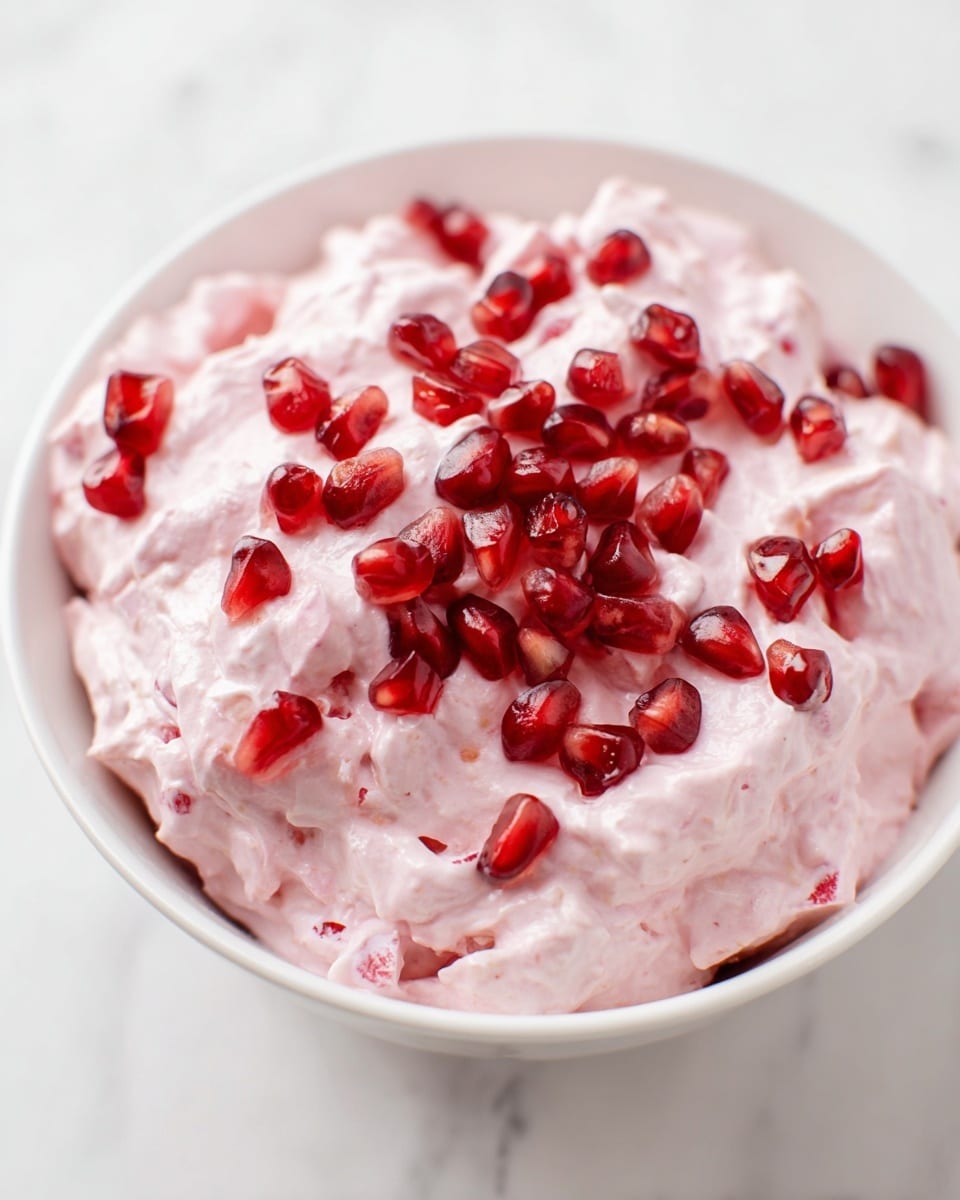 A white bowl filled with a pink creamy mixture that looks soft and fluffy, with small chunks visible inside. On top, there are many shiny, red pomegranate seeds scattered evenly, adding a bright contrast to the pale pink cream. The bowl sits on a white marbled surface, and the image is closely focused on the dessert, showing its smooth texture and vibrant seed details. photo taken with an iphone --ar 4:5 --v 7