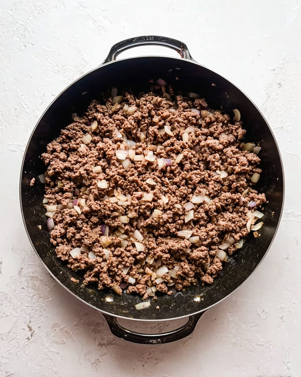 A top view of a large black pan with two handles, filled with cooked brown ground meat mixed with small white onion pieces. The meat is spread evenly, showing some browned bits and the onions are translucent and scattered throughout. The pan rests on a white marbled surface, creating a light, clean background. photo taken with an iphone --ar 4:5 --v 7