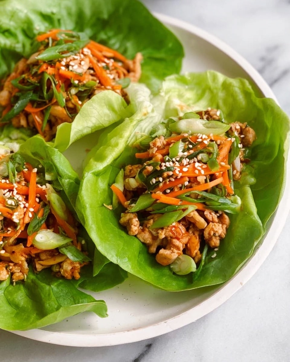 The image shows three green lettuce leaves used as cups, each filled with a colorful mix. The bottom layer is a large, smooth, bright green lettuce leaf. On top, there is a mix of light brown pieces of cooked food combined with thin, orange carrot strips and small green onion slices. White sesame seeds are sprinkled over the mix. The lettuce cups rest on a white plate placed on a white marbled surface. The colors are fresh and clear, showing textures of leafy, soft, and slightly crunchy ingredients. Photo taken with an iphone --ar 4:5 --v 7