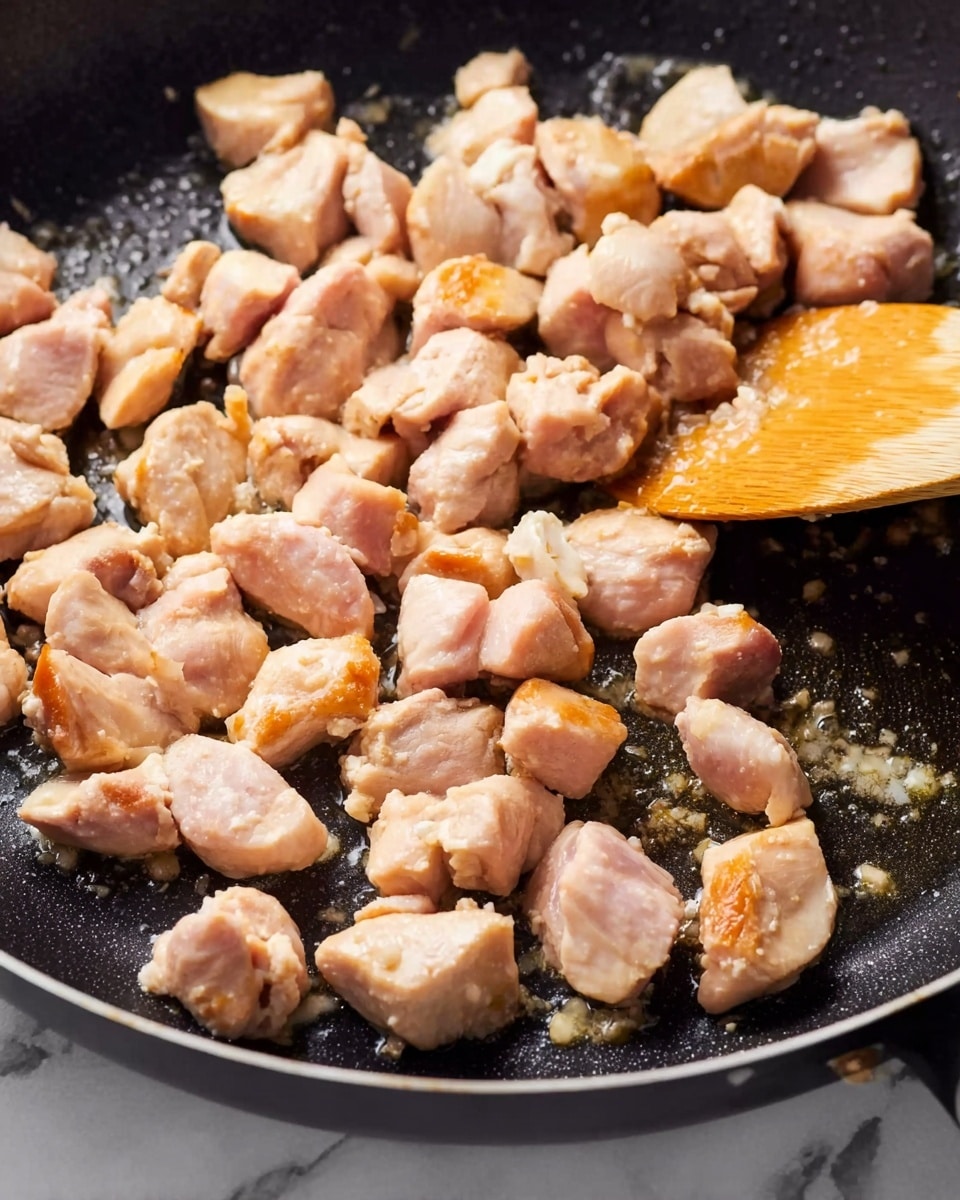 Close-up of small, uneven pieces of chicken cooking in a black frying pan. The chicken pieces are light pink with some golden brown areas showing they are fried on one side. A wooden spoon is stirring the chicken on the right side of the pan. The frying pan surface is slightly shiny with oil, and the background shows a white marbled texture. Photo taken with an iphone --ar 4:5 --v 7