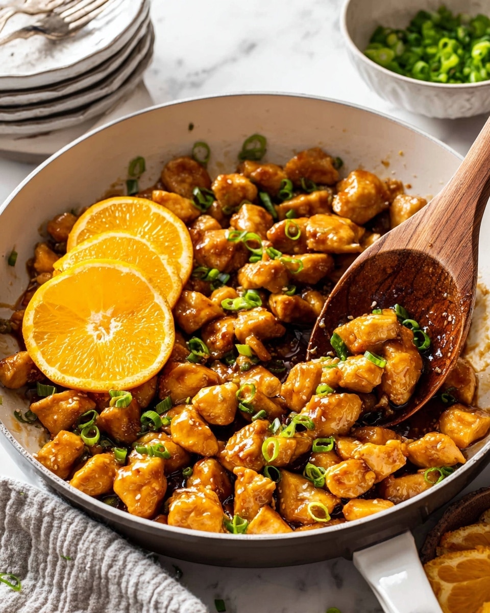 The image shows a white skillet filled with small, golden-brown chicken pieces coated in a shiny sauce. The chicken is mixed with green onion slices scattered on top, adding a pop of green color. On top in the center, there are two thin orange slices that add brightness to the dish. A wooden spoon lifts some of the chicken on the right side, showing more of the juicy texture. In the background, there are stacked white plates and a bowl with more green onion slices on a white marbled surface. Photo taken with an iphone --ar 4:5 --v 7