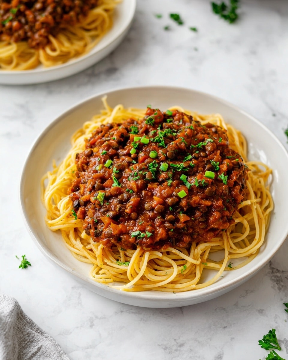 A white round plate on a white marbled surface holds a serving of spaghetti, forming the bottom layer with a slightly twisted, smooth yellow texture. On top of the spaghetti lies a thick layer of rich brownish-red sauce mixed with lentils and small bits of vegetables, adding a chunky texture. The sauce is garnished with small pieces of bright green chopped parsley scattered evenly over the top. In the background, part of another similar plate with the same dish is visible. Photo taken with an iphone --ar 4:5 --v 7