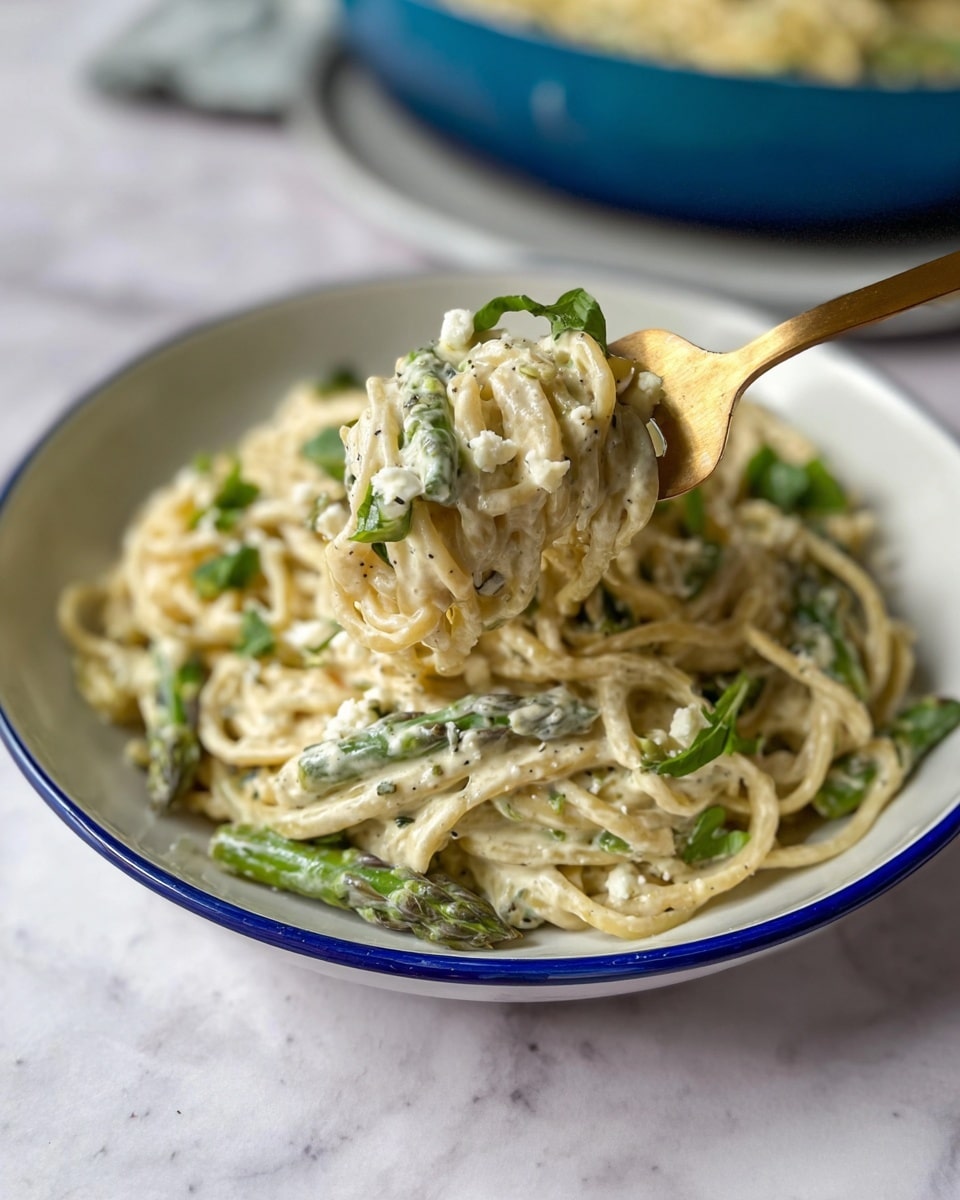 A white bowl with a thin blue rim holds creamy pasta with visible asparagus pieces mixed in. On top of the pasta, some green herbs and small white cheese bits add texture. A gold fork holds a close-up portion of the pasta, showing creamy white sauce coating the noodles and more herbs. The bowl sits on a white marbled surface, and in the background, there is a blurry blue dish with more pasta. photo taken with an iphone --ar 4:5 --v 7