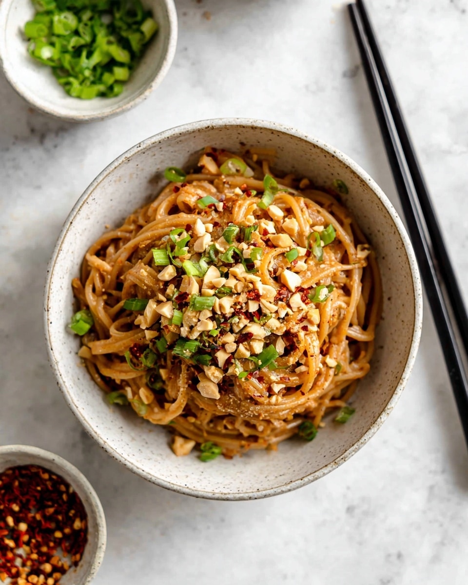 A white speckled bowl filled with a serving of brown noodles, coated in a light sauce, topped with a scattering of chopped green onions and crushed peanuts, and sprinkled with red chili flakes for a touch of color and texture. The bowl sits on a white marbled surface, with two black chopsticks placed diagonally next to it. In the background, two small white bowls hold more chopped green onions and chili flakes, adding subtle hints of green and red to the scene. Photo taken with an iphone --ar 4:5 --v 7