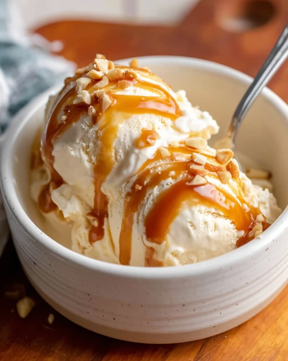 The image shows a white ceramic bowl filled with two scoops of creamy vanilla ice cream that look smooth and soft. The ice cream is topped with a shiny golden caramel sauce drizzled evenly over it, creating thin, flowing lines that drip slightly down the sides of the scoops. There are small bits of nuts scattered on top, adding a crunchy texture to the sweet dessert. A silver spoon is placed inside the bowl, slightly inserted into the ice cream on the right side. The bowl is set on a wooden surface with a blurred background. Photo taken with an iphone --ar 4:5 --v 7
