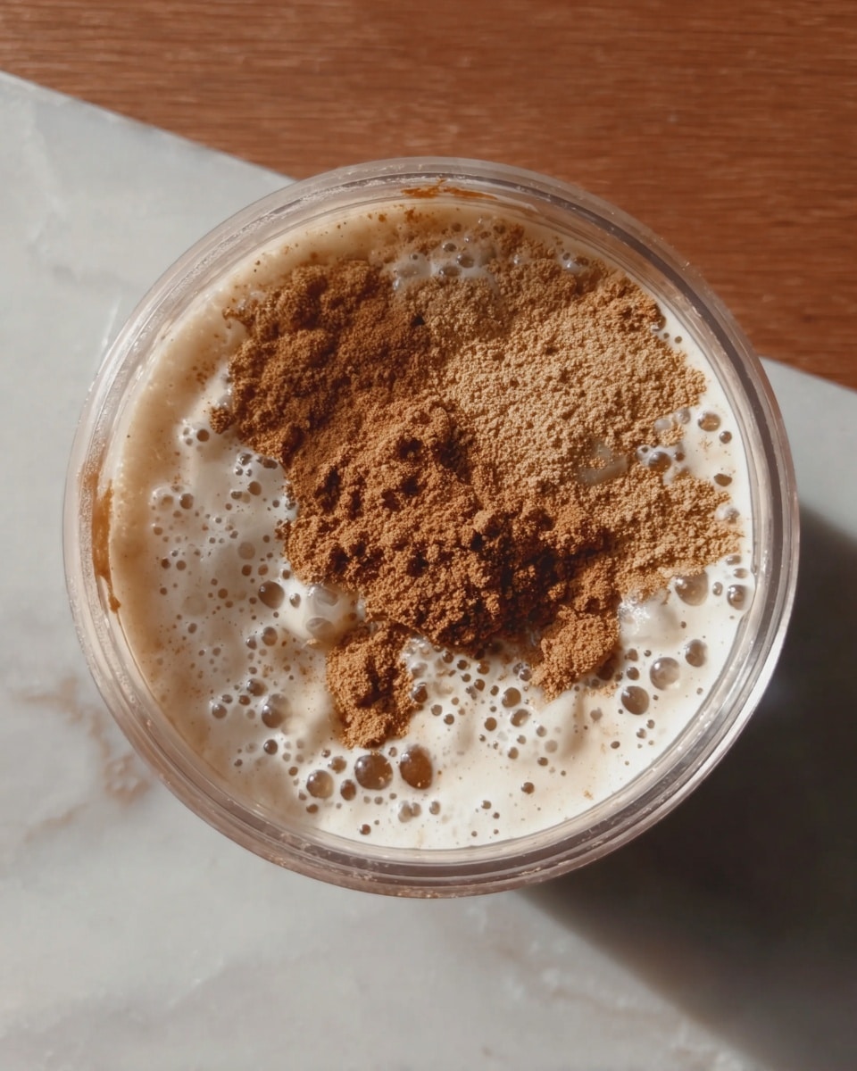 A clear round container filled with a creamy white liquid that has small bubbles on the surface, topped with a heap of light brown powder and some darker brown powder sprinkled unevenly in the center. The container is placed on a white marbled surface. photo taken with an iphone --ar 4:5 --v 7