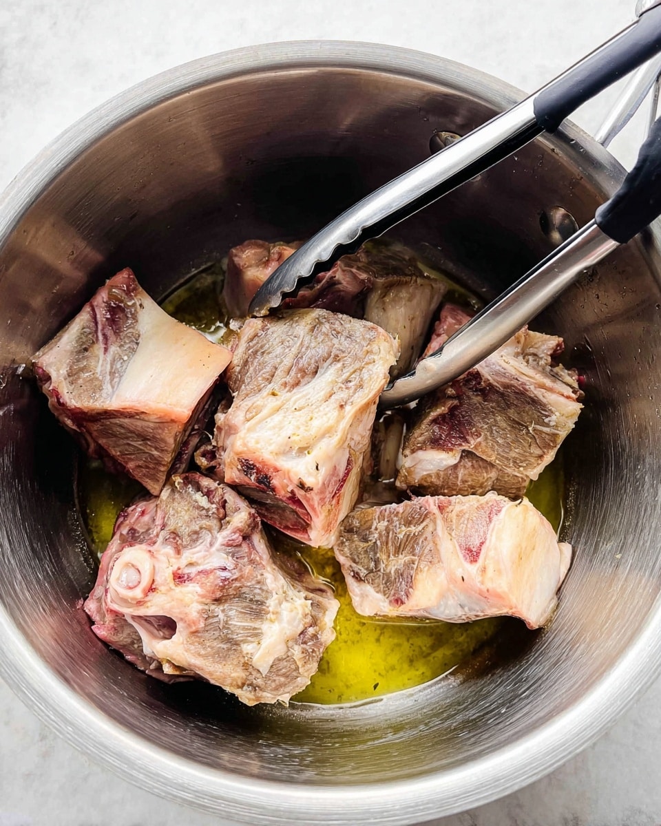 Inside a large silver metal pot are seven pieces of meat with bones, showing a mix of light pink, pale white, and some brown seared parts. The meat pieces have different shapes and sizes, some with visible bone edges. They sit in a small amount of yellowish oil at the bottom of the pot. A pair of black kitchen tongs with silver handles hold one piece of meat near the center. The background surface is a white marbled texture. photo taken with an iphone --ar 4:5 --v 7