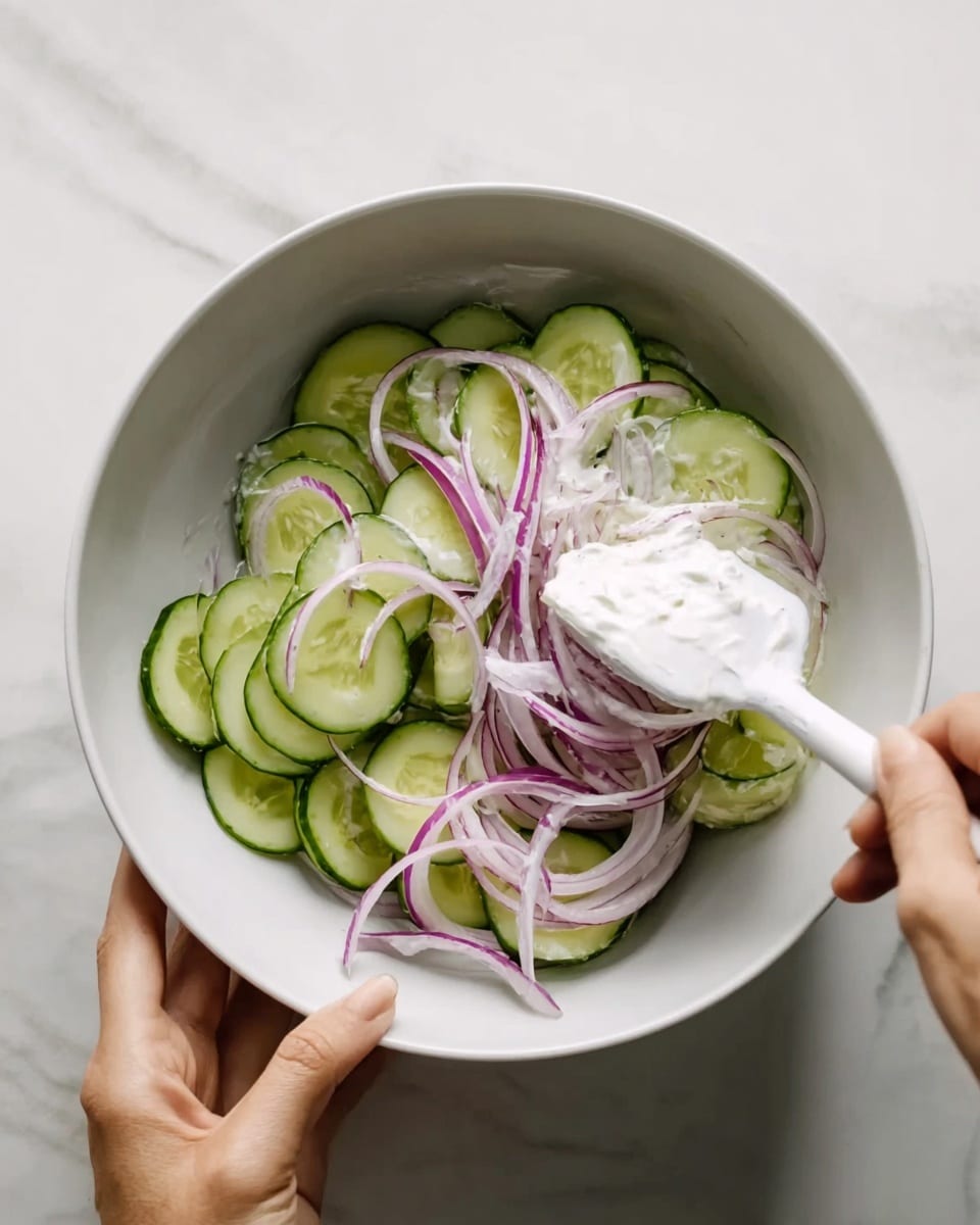 A white bowl holds thin, round slices of green cucumber forming the bottom layer, topped with thin wedges of purple onion arranged around the edges. A creamy white dressing is being mixed into the vegetables with a white spatula held by a woman's hand, while another woman's hand steadies the bowl against a white marbled surface. The textures contrast between the smooth cream, the fresh, crisp cucumber, and the slightly papery onion slices. photo taken with an iphone --ar 4:5 --v 7