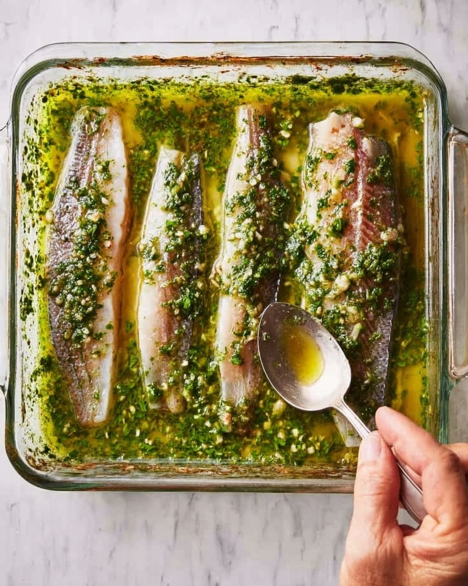 The image shows a clear glass rectangular dish with three split fish fillets placed on the left side and one whole fish fillet on the right, all lying in a thick green herb sauce mixed with garlic and small bits of seasoning. The green sauce has a slightly chunky texture with visible pieces of herbs and garlic scattered throughout. A woman's hand is holding the whole fish fillet on the right, while a spoon rests in the sauce near the top right corner of the dish. The dish is placed on a white marbled surface. photo taken with an iphone --ar 4:5 --v 7